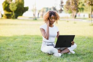 Young attractive dark-skinned college student wearing tank top and jeans sitting on the lawn at campus on sunny day, working on her thesis using laptop computer, looking busy and concentrated photo