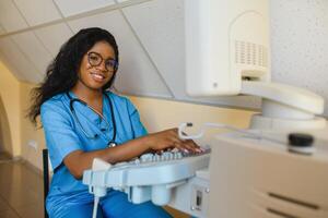 Smiling African woman doctor with ultrasound scanner in hand, working on modern ultrasound scanning machine in light room in clinic. Portrait Of 4D Ultrasound Scanning Machine Operator photo