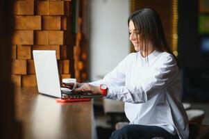 joven mujer exitoso gerente es hablando vía móvil teléfono con posible financiero empresa empleado y leyendo su currículum en portátil ordenador portátil computadora durante remoto entrevista en trabajo colaborativo espacio foto