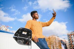 eléctrico carros, ev concepto, eco simpático combustible. retrato de joven sonriente negro hombre, recarga su moderno lujo eléctrico coche foto