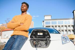 eléctrico carros, ev concepto, eco simpático combustible. retrato de joven sonriente negro hombre, recarga su moderno lujo eléctrico coche foto