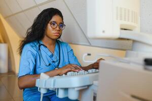 Young african-american female doctor in white coat using ultra ultrasound scanning machine and looking on the screen. African woman doctor working on modern ultrasound equipment. photo