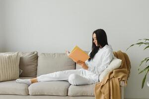 bonito joven mujer disfrutando leyendo un libro a hogar acostado en el sofá sonriente en Placer en casual ropa. foto