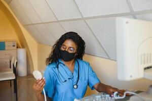 joven alegre africano mujer operador en máscara de un ultrasonido exploración máquina analizando diagnósticos resultados de paciente. joven sonriente africano médico trabajando en un moderno ultrasonido equipo. foto