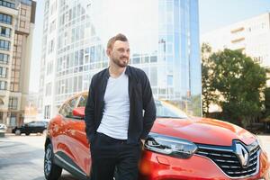 un atractivo hombre con un barba soportes siguiente a su coche en el estacionamiento lote. él es considerado, y mira lejos, alguien es espera. verano día foto