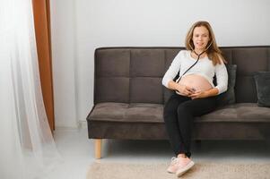 Pregnant woman using stethoscope examining her belly on sofa, Happy pregnant woman Concept. photo