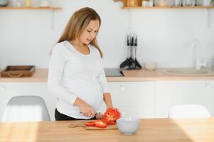 pregnancy, cooking food, healthy lifestyle, people and expectation concept - happy pregnant woman with knife chopping cucumber and preparing vegetable salad at home photo