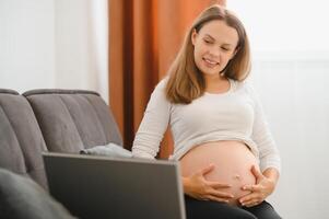 Pregnant woman video chatting with family on laptop waving hand to screen photo