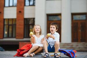 primary education, friendship, childhood and people concept - group of happy elementary school students with backpacks. Two schoolchildren sitting on books near school and eating an apple. photo