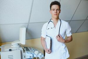 encantador joven masculino médico , sentado a su oficina cerca ultrasonido exploración máquina. hermoso simpático médico disfrutando trabajando a el hospital, Copiar espacio. ultrasónico concepto foto