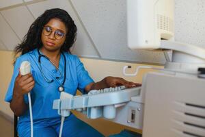 Young female African-american doctor working on modern ultrasound equipment. Operator of ultrasound scanning machine sitting and looking at the monitor, waiting for patient. photo