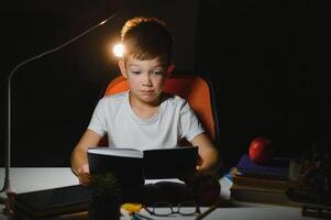 concentrated schoolboy reading book at table with books, plant, lamp, colour pencils, apple, and textbook photo