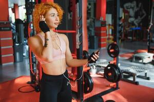 Smiling charming young woman athlete in earphones using smartphone and listening to music in gym photo