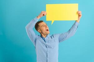 Handsome smiling man holding blank speech bubble sign isolated on light blue studio banner background with copy space photo