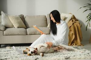 joven sonriente mujer descansando en sofá y leyendo un libro con perro sentado cerca por su a hogar. foto