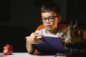 concentrated schoolboy reading book at table with books, plant, lamp, colour pencils, apple, and textbook photo