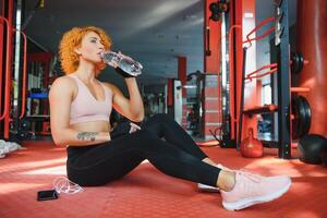hermosa joven deporte mujer cansado tomando un descanso desde ejercicio sentado en piso Bebiendo agua y toalla sudor y pesa en aptitud gimnasio .chica en ropa de deporte rutina de ejercicio descanso foto