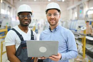 Two Diverse Professional Heavy Industry Engineers Wearing Safety Uniform and Hard Hats Working on Laptop photo