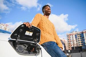Electric cars, EV concept, eco friendly fuel. Portrait of young smiling black man, recharging his modern luxury electric car photo