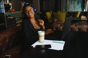 Happy black girl in wireless headphones studying online, using laptop and taking notes, cafe interior, copy space photo