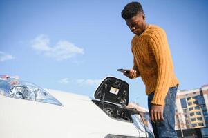 Close up of african american man connecting charging cable to electric car. Young male standing near his modern auto with leather suitcase in hand. photo