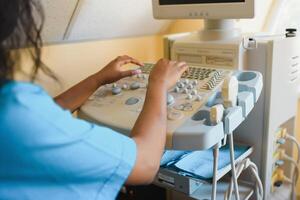 Young female African-american doctor working on modern ultrasound equipment. Operator of ultrasound scanning machine sitting and looking at the monitor, waiting for patient. photo