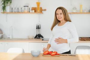 el embarazo, Cocinando alimento, sano estilo de vida, personas y expectativa concepto - contento embarazada mujer con cuchillo el cortar Pepino y preparando vegetal ensalada a hogar foto