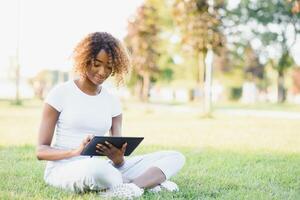 Thoughtful cute mixed female international student with curly hair is sitting on fresh grass with modern laptop in public park, leaning on apple tree and wistfully looking aside during her break photo