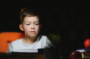 concentrated schoolboy reading book at table with books, plant, lamp, colour pencils, apple, and textbook photo