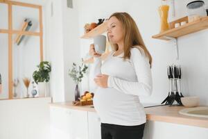 imagen de hermosa por casualidad vestido joven morena hembra esperando bebé, teniendo Mañana té en moderno elegante cocina, sonriente. anticipación, expectativa, parto y maternidad concepto foto