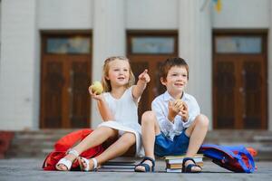 primary education, friendship, childhood and people concept - group of happy elementary school students with backpacks. Two schoolchildren sitting on books near school and eating an apple. photo