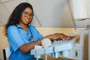 Young female African-american doctor working on modern ultrasound equipment. Operator of ultrasound scanning machine sitting and looking at the monitor, waiting for patient. photo