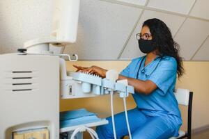 joven hembra afroamericano médico trabajando en moderno ultrasonido equipo. operador de ultrasonido exploración máquina sentado y mirando a el monitor, esperando para paciente. foto