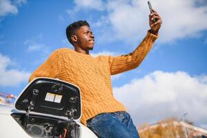 Portrait of young handsome black man in business suit, making selfie photo on his smartphone while leaning on his electric car, charging the battery at city power station