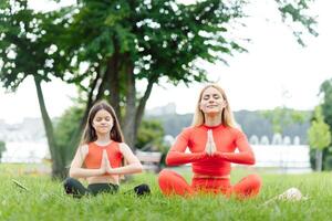 madre y hija haciendo yoga ejercicios en césped en el parque a el día tiempo. personas teniendo divertido al aire libre. concepto de simpático familia y de verano vacaciones. foto