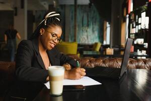 Student girl browsing Internet, using free wi-fi at cafe. African freelancer thinking on ideas for her blog, using laptop at co-working space, resting hand on wooden table, looking with inspired smile photo