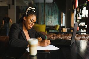 estudiante niña hojeada Internet, utilizando gratis Wifi a cafetería. africano persona de libre dedicación pensando en ideas para su Blog, utilizando ordenador portátil a trabajo colaborativo espacio, descansando mano en de madera mesa, mirando con inspirado sonrisa foto