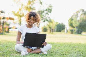 Thoughtful cute mixed female international student with curly hair is sitting on fresh grass with modern laptop in public park, leaning on apple tree and wistfully looking aside during her break photo