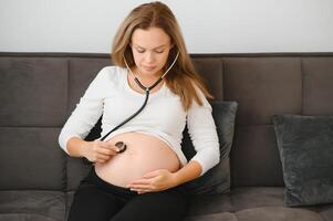 The mother is using stethoscope the doctor listens to the sound of the baby in the stomach. Asian pregnant woman using stethoscope listening her baby on sofa. photo