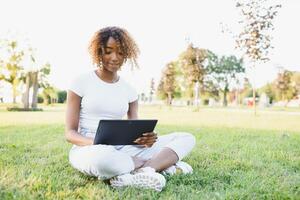 pensativo linda mezclado hembra internacional estudiante con Rizado pelo es sentado en Fresco césped con moderno ordenador portátil en público parque, propensión en manzana árbol y con nostalgia mirando aparte durante su descanso foto