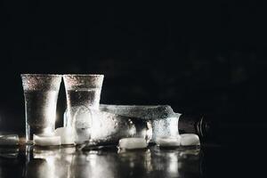 Close-up view of bottle of vodka with glasses standing on ice on black. photo