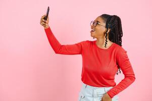 Smiling young african american woman girl in sweatshirt posing isolated on pastel pink wall background in studio. People lifestyle concept. Mock up copy space. Doing selfie shot on mobile phone. photo