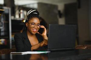 Young girl in glasses amazingly looking in laptop at cafe. African American girl sitting in restaurant with laptop and cup on table. Portrait of surprised lady with dark curly hair in earphones photo