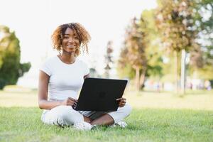 Young smiling black woman sitting outdoors on grass with laptop, typing, surfing internet. Technology, communication, education and remote working concept, copy space photo