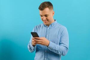 Photo of happy handsome man holding mobile phone in hand, texting. Young guy with big toothy smile posing on blue background.