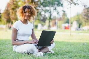 pensativo linda mezclado hembra internacional estudiante con Rizado pelo es sentado en Fresco césped con moderno ordenador portátil en público parque, propensión en manzana árbol y con nostalgia mirando aparte durante su descanso foto