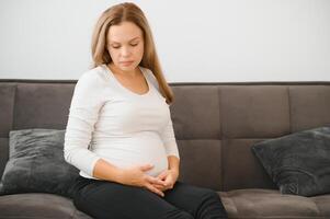 hermosa embarazada mujer sentado a sofá y acuerdo mano en barriga. foto
