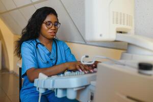 Smiling African woman doctor with ultrasound scanner in hand, working on modern ultrasound scanning machine in light room in clinic. Portrait Of 4D Ultrasound Scanning Machine Operator photo