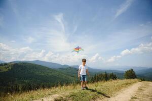 Little school age boy running down the slope with kite in the sun. Sunny summer or spring day at sunset. Active outdoor games and leisure. Mountain landscape with hills and coniferous trees. photo