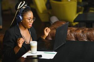 Happy black girl in wireless headphones studying online, using laptop and taking notes, cafe interior, copy space photo